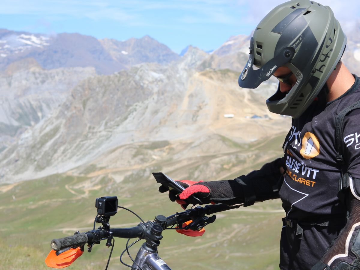 Primo piano su un supporto bici Shapeheart fissato al manubrio di una MTB, smartphone visibile, anche su terreno accidentato.