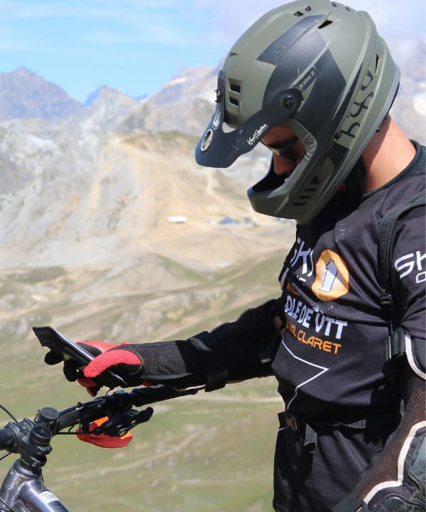 Primo piano su un supporto bici Shapeheart fissato al manubrio di una MTB, smartphone visibile, anche su terreno accidentato.